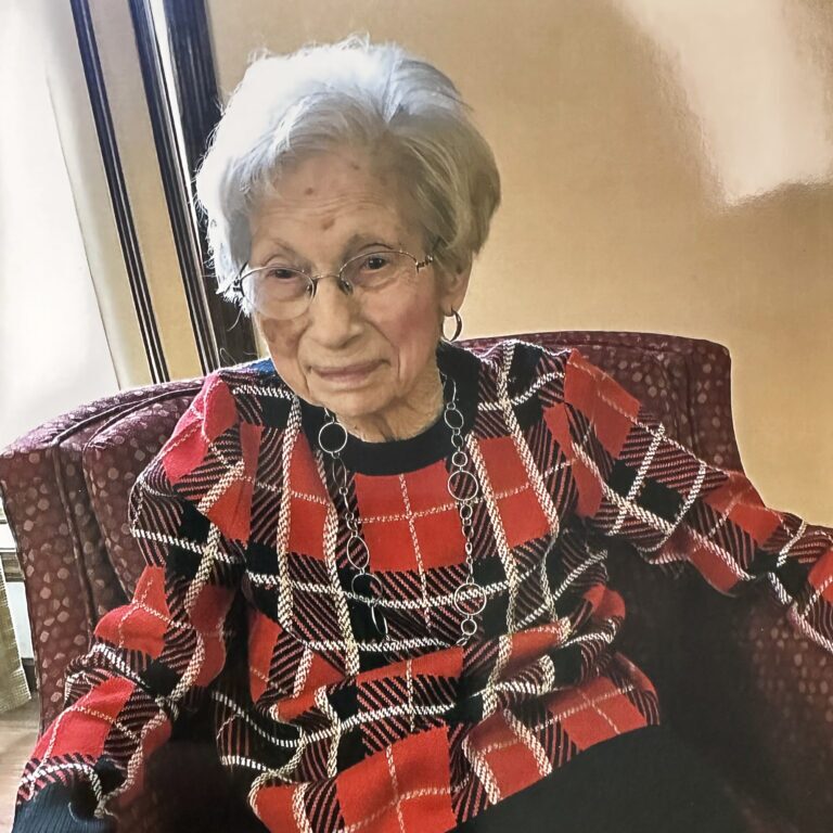 Centenarian Betty Whiteleather sitting in a chair while wearing a red and black plaid blouse