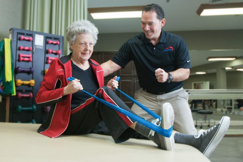 Senior woman with exercise band helped by male physical therapist.