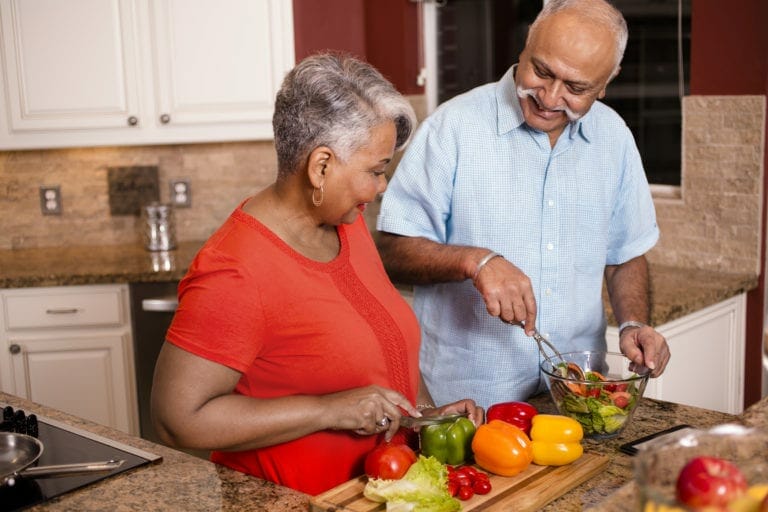 Senior adult (Indian and African descent) couple cooking dinner together at home. They are enjoying their retirement years as they work together to chop healthy vegetables for a salad.